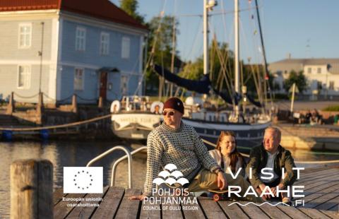 Kolme henkilöä istumassa laiturilla museon edustalla aurinkoisena kesäpäivänä.