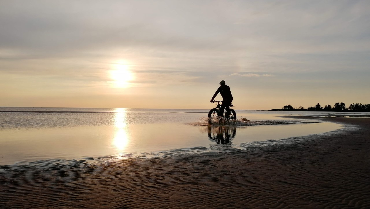 Pyöräilijä rannalla auringon laskiessa.
