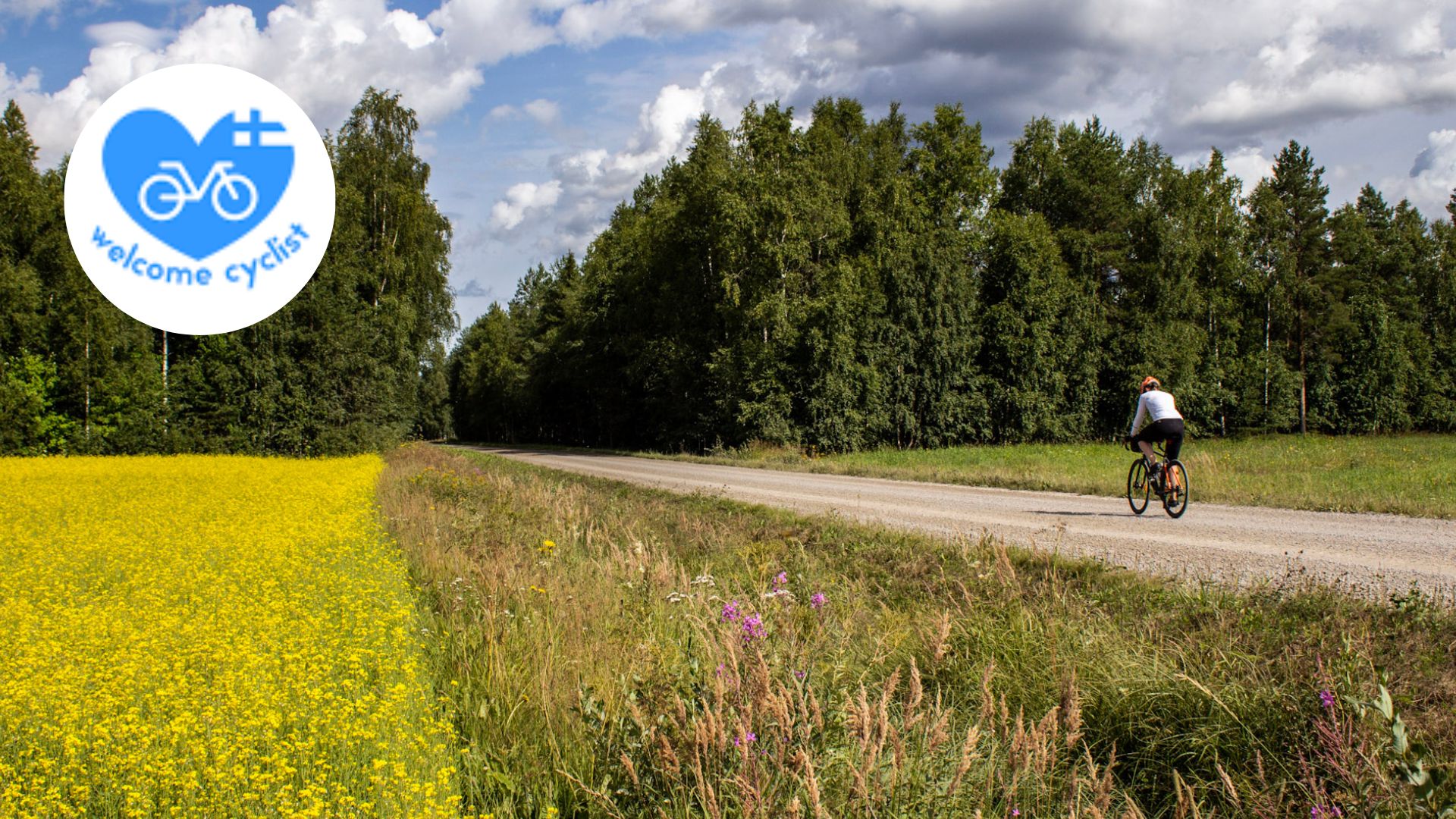 Pyöräilijä soratiellä ohittamassa pellot ja menossa metsään.